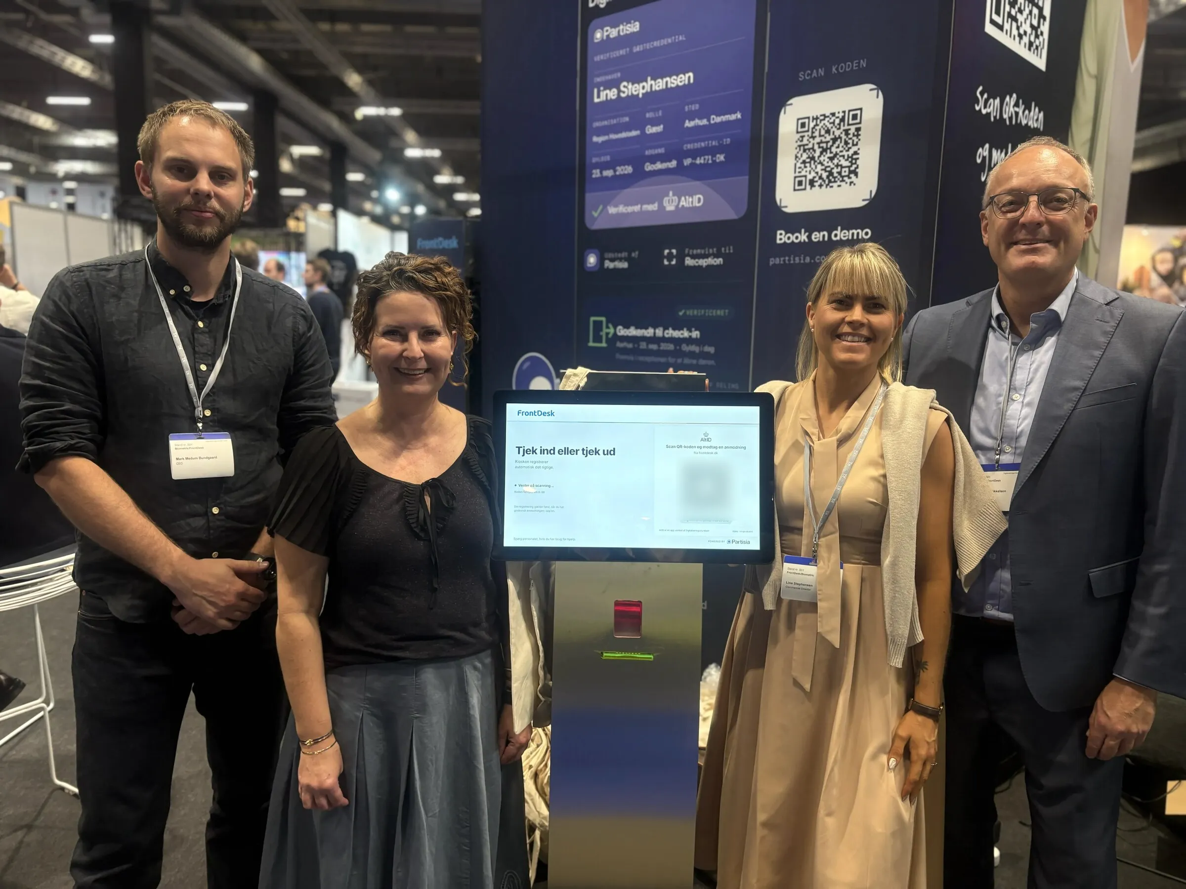 Four people standing either side of a FrontDesk self-service kiosk on a trade-fair stand. The kiosk screen shows the AltID check-in step, and the banner behind shows a verified guest credential.