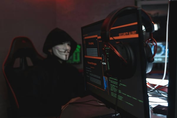 Person in a dark hoodie and Guy Fawkes mask sitting at a desk with multiple monitors displaying cybersecurity alerts and code, suggesting hacking activity.