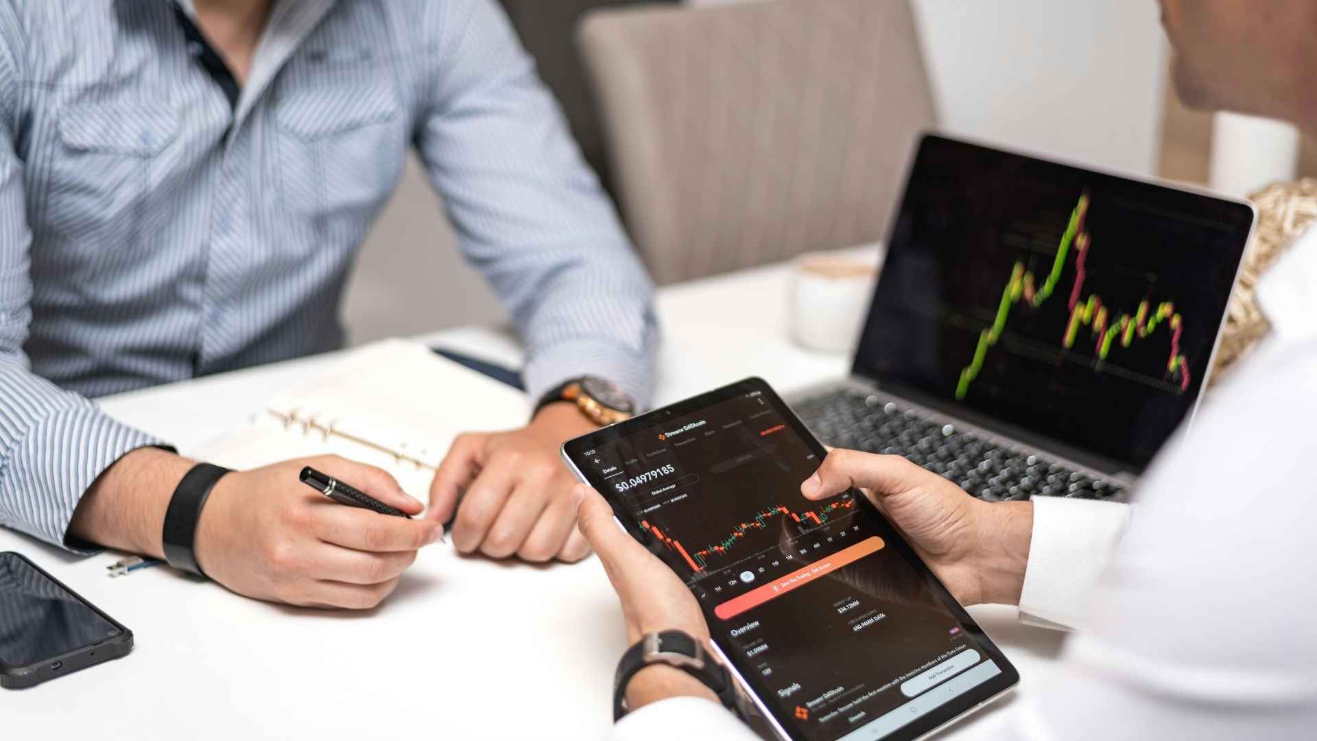 Two professionals analyzing financial data on a tablet and laptop, discussing fintech solutions for trading and investment.